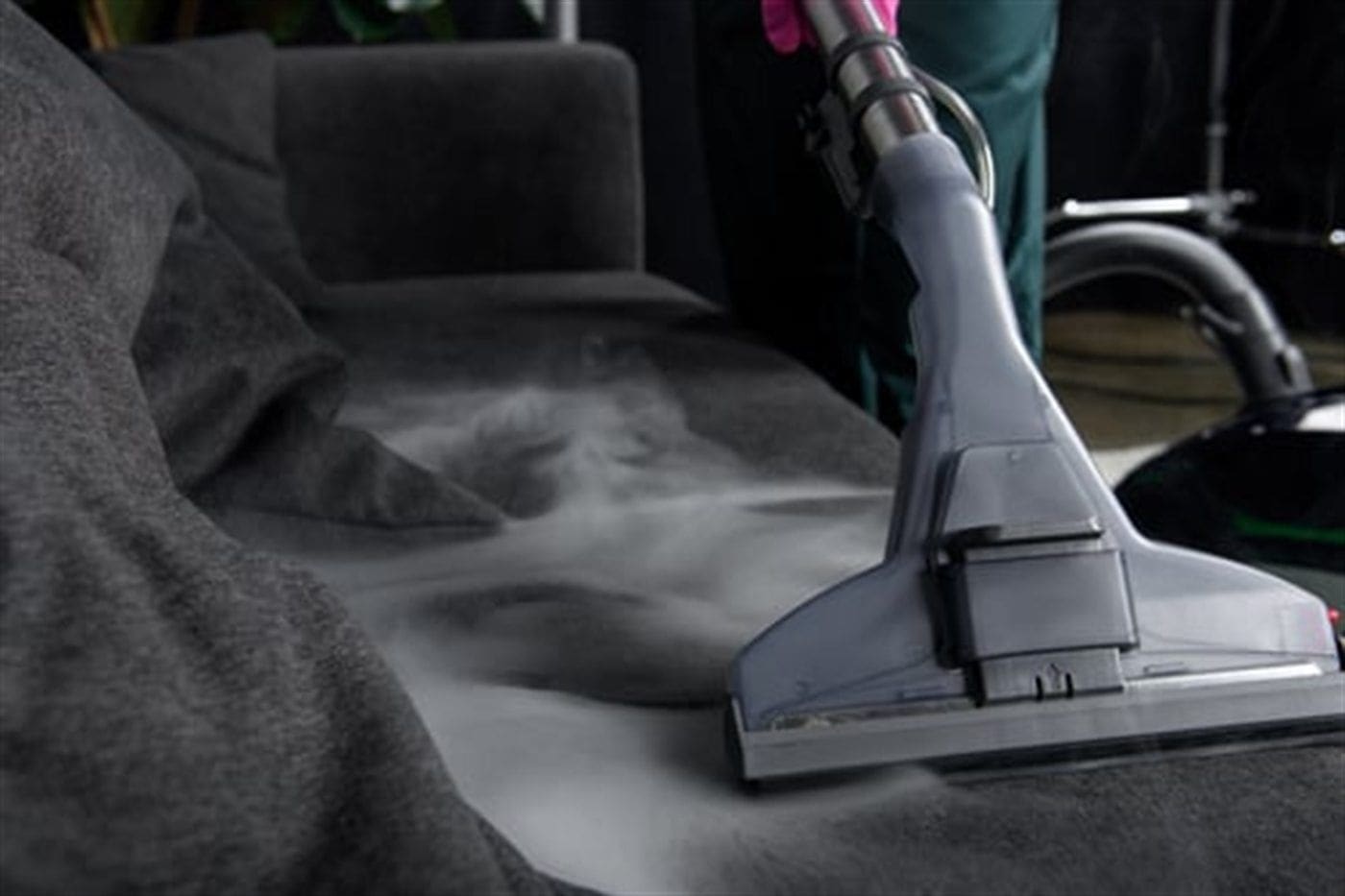 Upholstery steam cleaning wand lifting embedded soil from a dark fabric sofa with visible steam